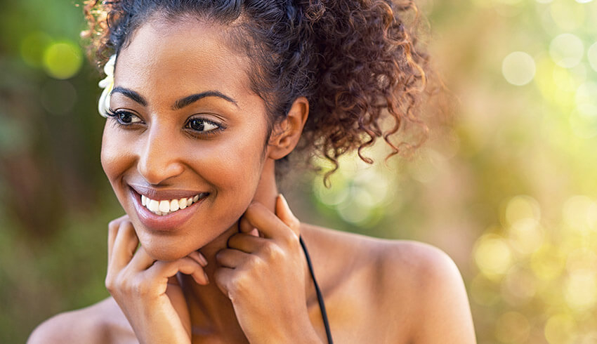 A smiling woman stands outside with her closed hands placed under her face.